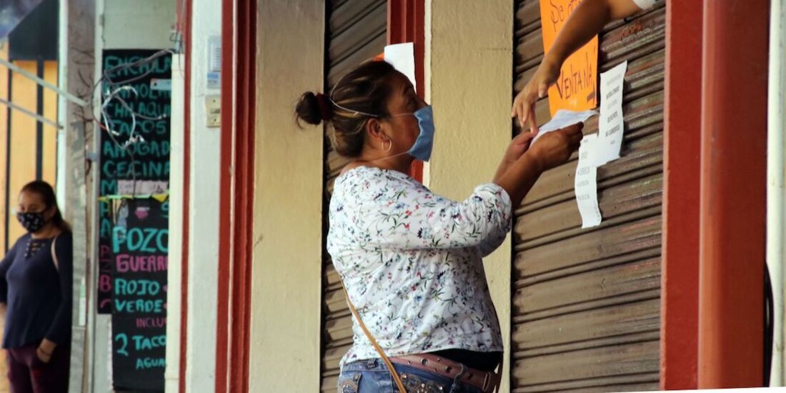 Una mujer acude a una farmacia a comprar, donde una trabajadora la atiende detrás de la cortina cerrada del negocio.