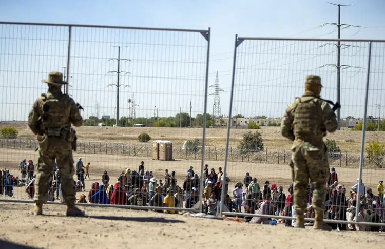 Migrantes esperan junto a una valla fronteriza mientras son vigilados por miembros de la Guardia Nacional de Texas, para ingresar a El Paso, Texas, el miércoles 10 de mayo de 2023.