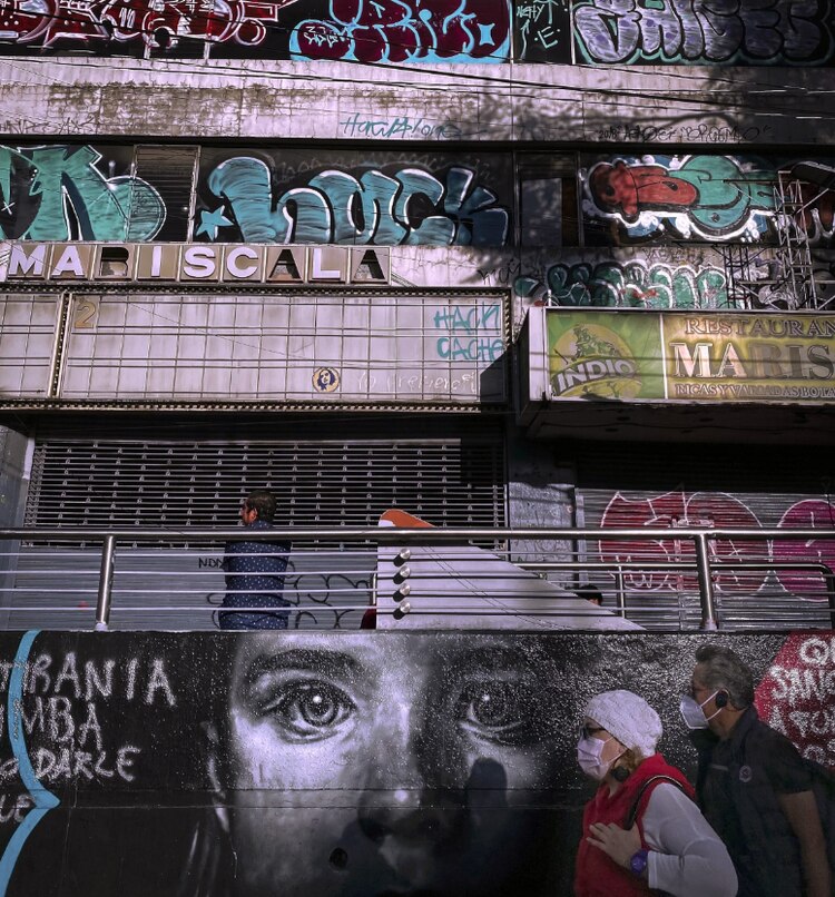 Un hombre y una mujer con cubrebocas pasan frente a un mural