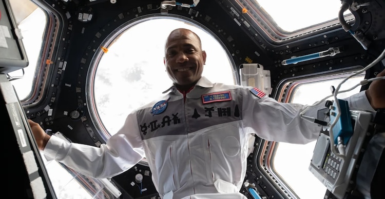 El ingeniero Victor J. Glover vestido con un traje de vuelo blanco frente a las ventanas del módulo Cupola
