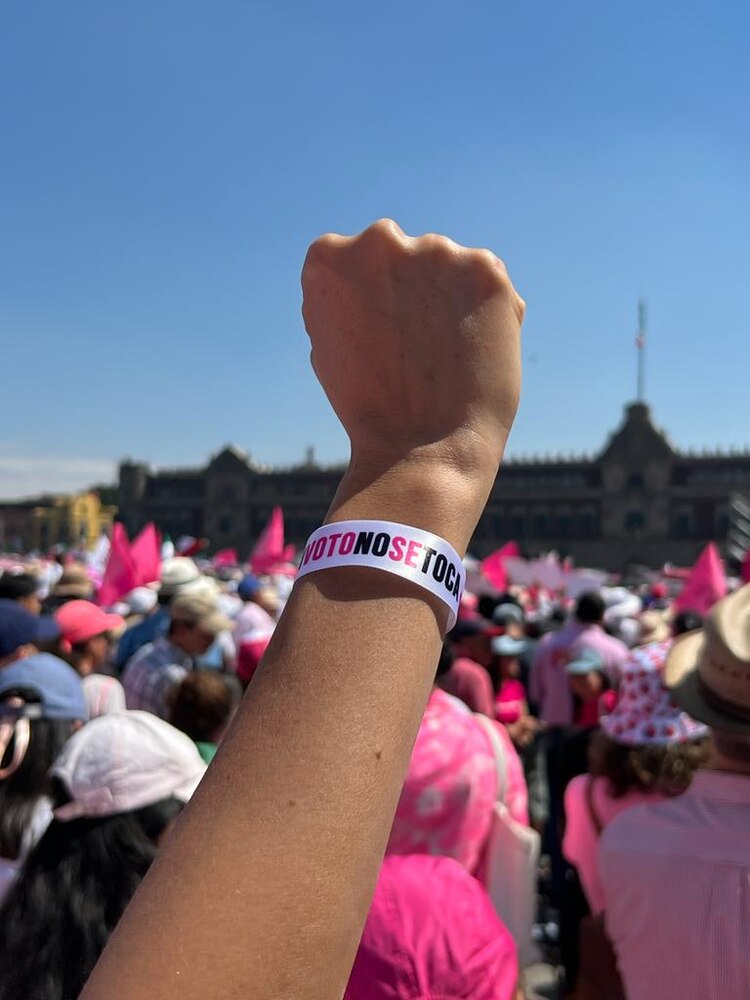 Durante la entonación del Himno Nacional de México, una mujer levanta su brazo; en su muñeca porta una pulsera con el lema: "Mi voto no se toca"