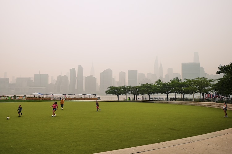 Un grupo de personas juega en el Hunter's Point South Park Athletic Field. Al fondo se aprecian los rascacielos de Nueva York, ocultos parcialmente por el humo de los incendios forestales en Canadá
