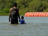 Migrantes caminan junto a grandes boyas que se utilizan como barrera fronteriza flotante en el río Bravo, el 1 de agosto de 2023, en Eagle Pass, Texas