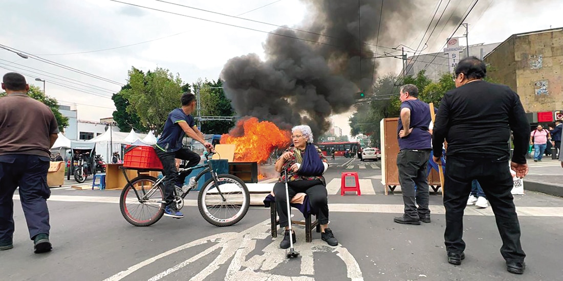 Una adulta mayor está sentada sobre Eje Central, mientras arden muebles, ayer.